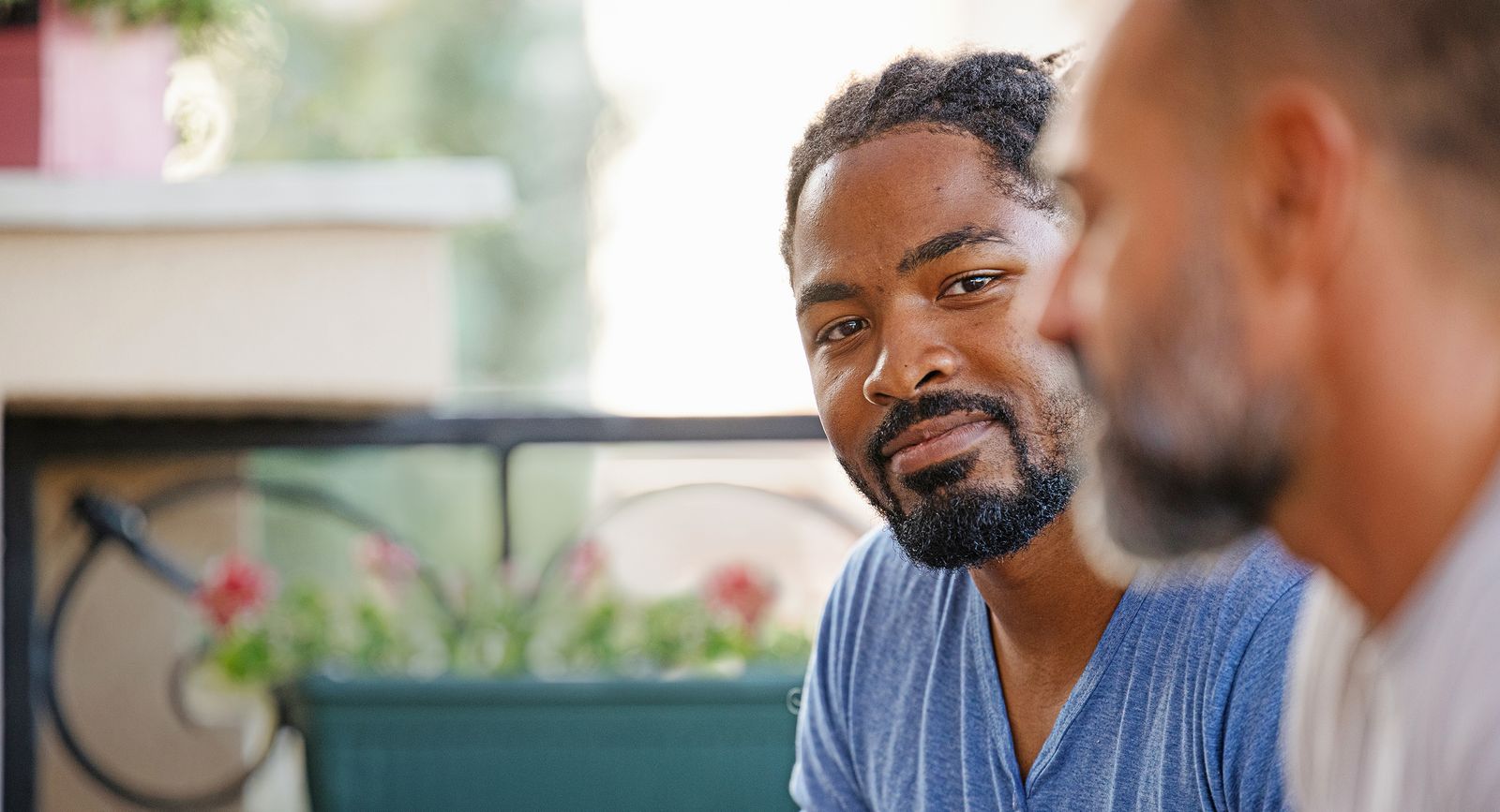 Two men are engaged in a conversation outdoors. The man in the foreground has a beard and is wearing a blue shirt, while the other man's face is partially visible. 