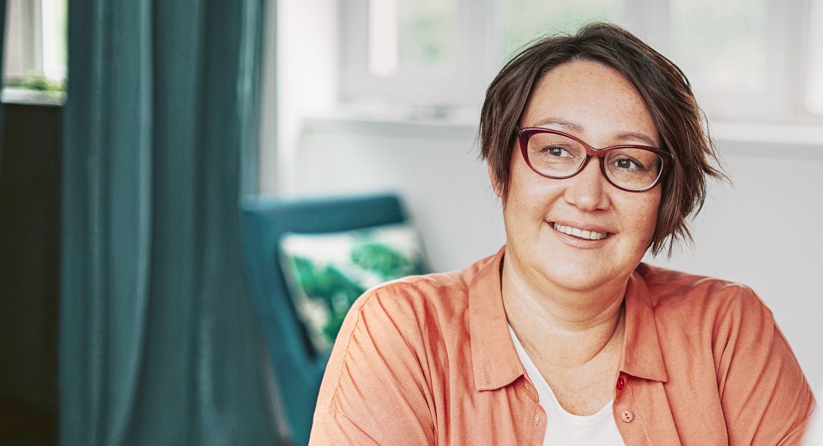 A woman with short hair and glasses, wearing an orange shirt, smiles while looking off-camera indoors.