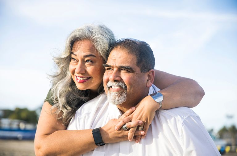 A woman with grey hair hugs a smiling man. Both are outdoors on a sunny day.