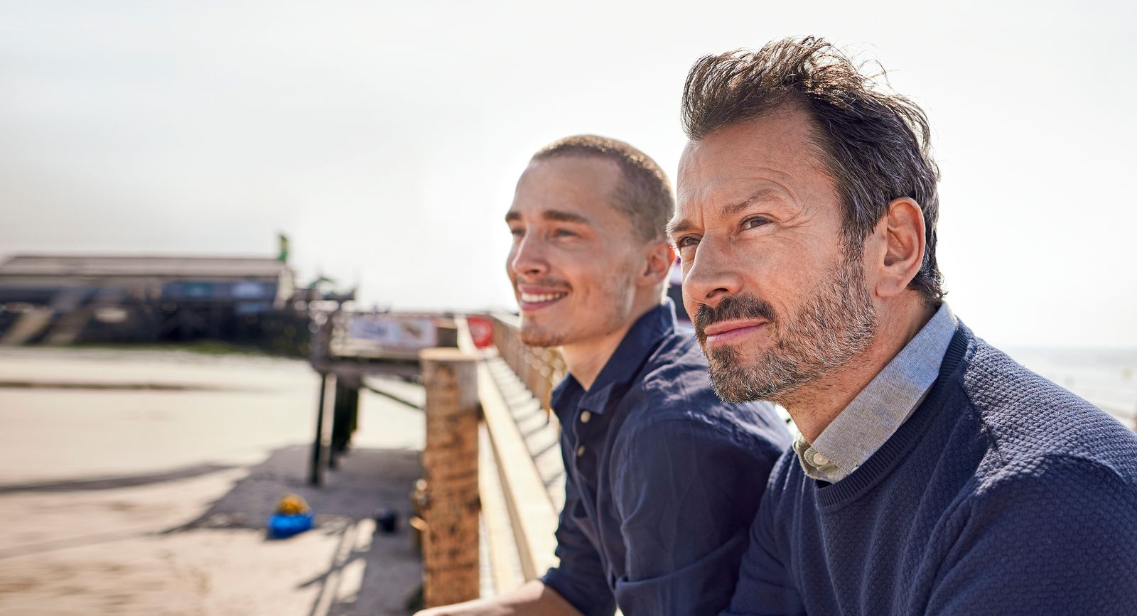 Two men sitting side by side on a seaside boardwalk, looking into the distance with a beach and pier structure in the background.