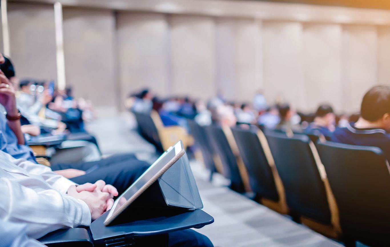 People seated in an auditorium, focused on a presentation, with notebooks and tablets in use. Rows of chairs are visible, and the setting appears to be a conference or lecture.