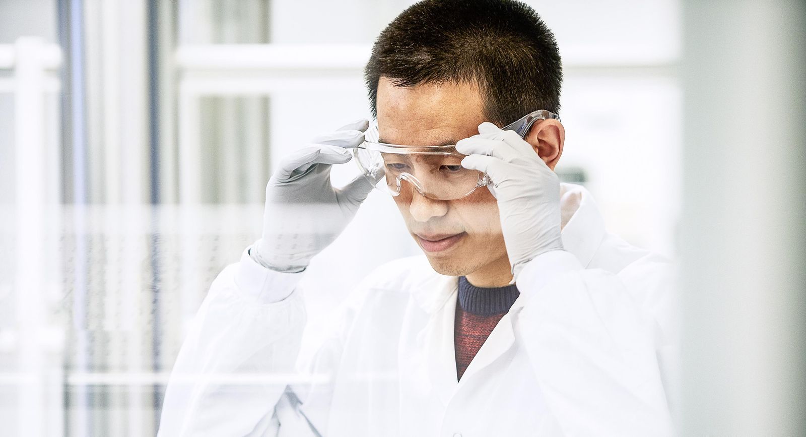 A Camurus' employee wearing a white lab coat, gloves, and safety goggles adjusts their eyewear while working in the laboratory