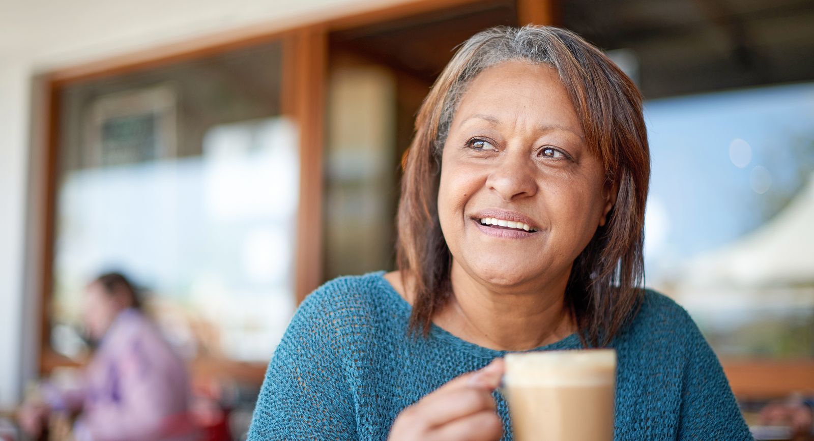 A woman smiles while holding a cup of coffee in an outdoor café setting.