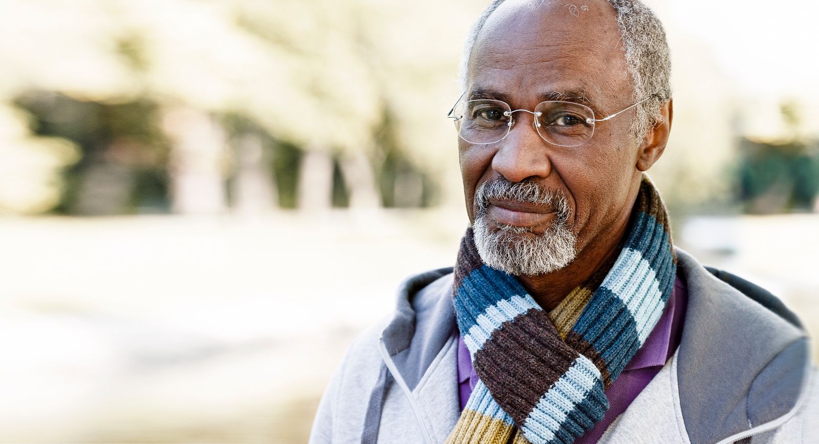 An elderly man is outdoors, wearing glasses, a jacket, and a striped scarf. He has a gray beard and short hair, and a contemplative expression. The background is blurred greenery.