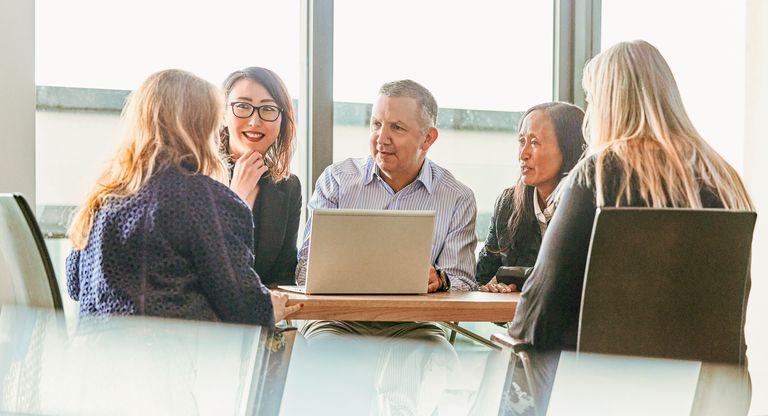 Camurus US employees sit around a table with a laptop, engaged in discussion. Natural light comes through large windows behind them.