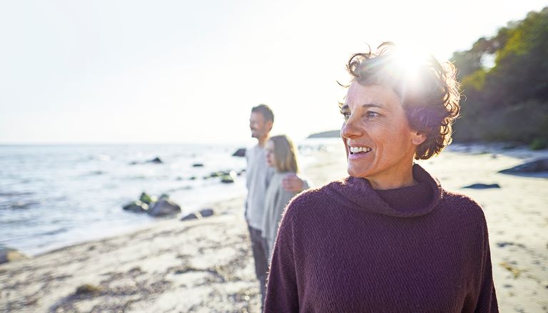 A person wearing a purple sweater smiles while standing on a beach. In the background, a man and a child look out at the water.