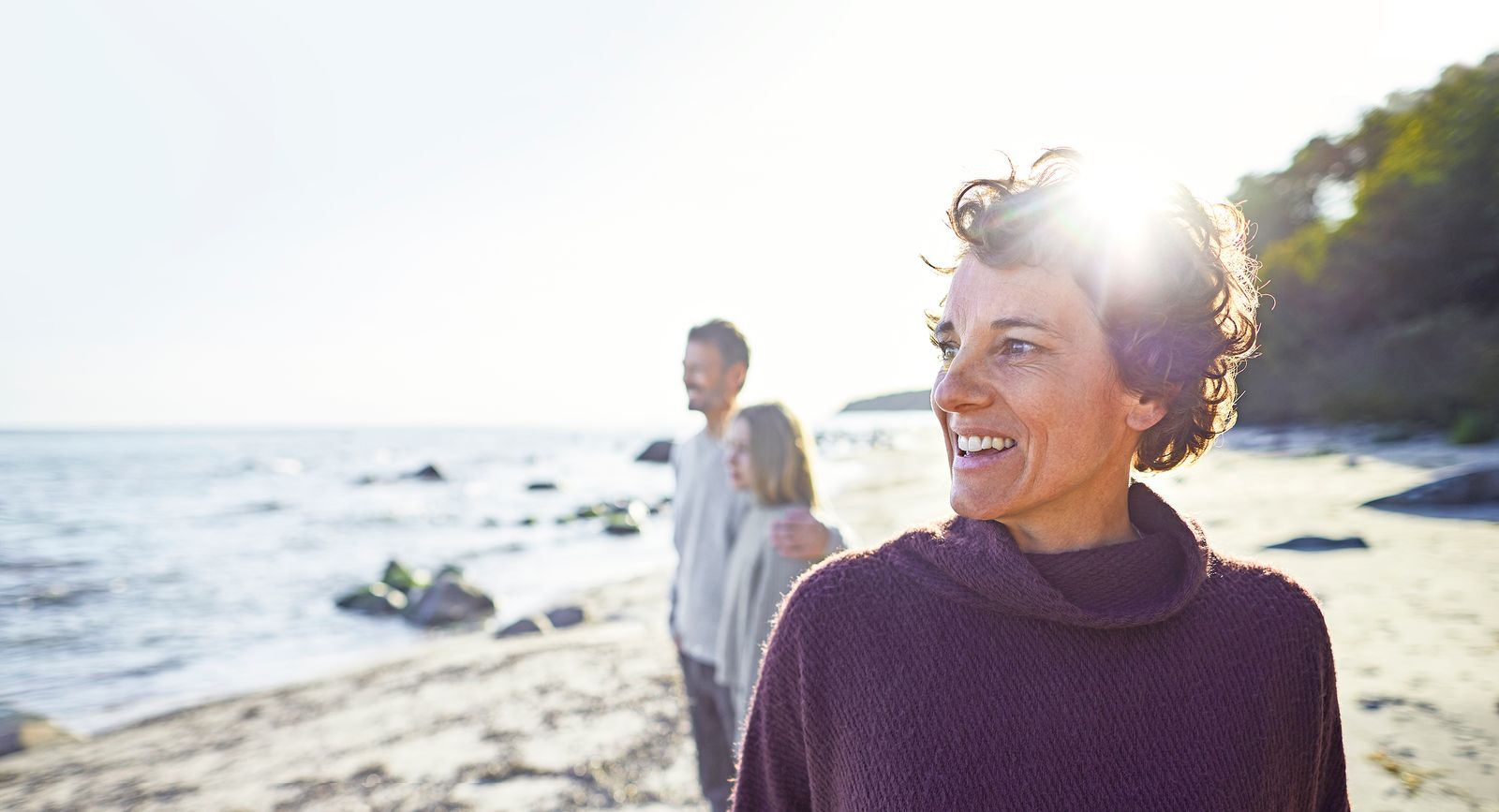 A person wearing a purple sweater smiles while standing on a beach. In the background, a man and a child look out at the water.